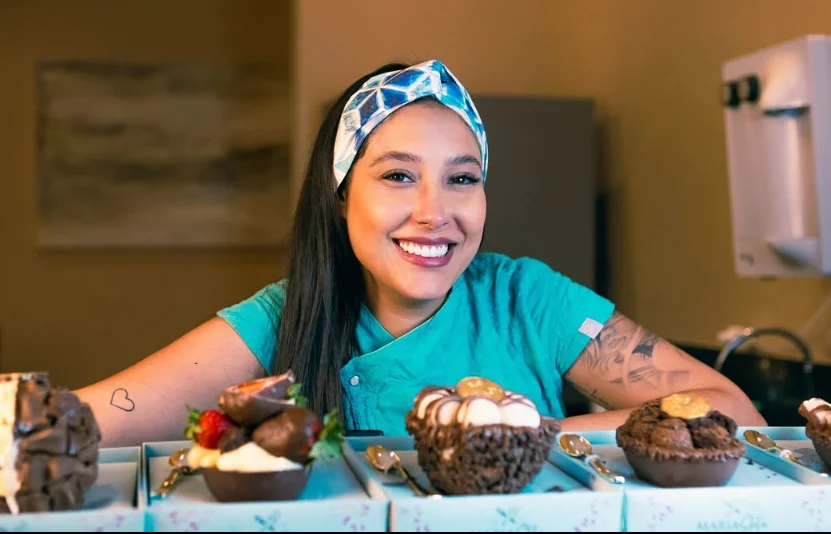 Maria Stela sorrindo na cozinha, atrás de doces preparados por ela